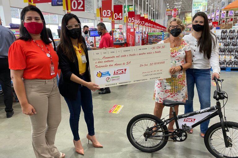 A senhora Alice Kuerten, da Federação das APAEs, recebe o cheque do Troco Solidário de Fernanda Tambellini, Priscila Santana e Bianca Pinheiro do Fort Atacadista